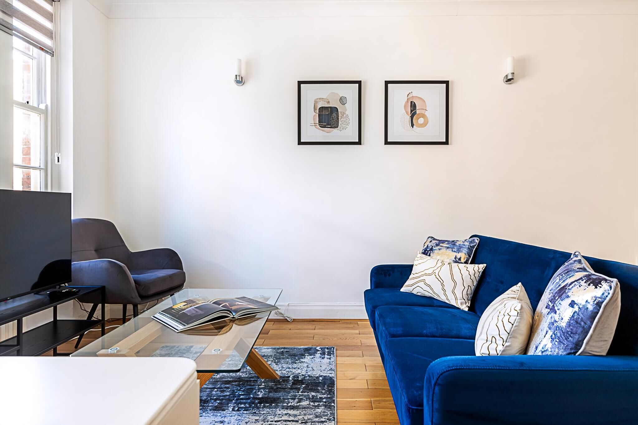 Ein modernes Wohnzimmer mit einem blauen Sofa, zwei Sesseln, einem Couchtisch und einem Fernseher. An der Wand befinden sich zwei gerahmte Kunstwerke.