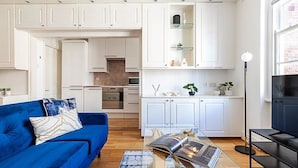 Un salon moderne avec un canapé bleu, une table basse sur laquelle repose un magazine, et un coin cuisine doté de meubles blancs et d’un lampadaire noir.