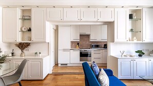 Une cuisine moderne avec des meubles blancs, un coin repas doté d’une table et de chaises en verre, et un canapé bleu agrémenté de coussins décoratifs.