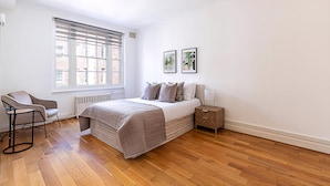 Une chambre à coucher comprenant un lit, une chaise, une table de chevet, une fenêtre avec des stores et deux tableaux encadrés accrochés au mur.
