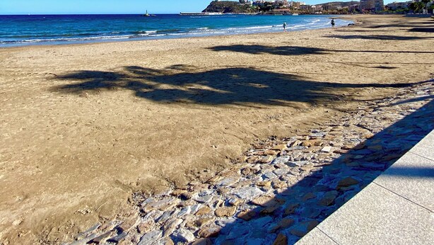 Una playa con cielos despejados, costa rocosa y edificios a lo lejos.