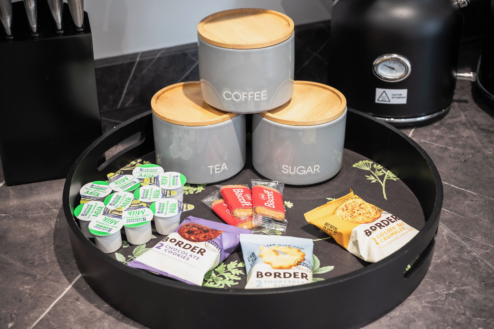 Un plan de travail de cuisine sur lequel se trouvent trois récipients étiquetés « café », « thé » et « sucre ». On y trouve également divers en-cas et condiments emballés.