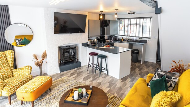 A modern living room with a sofa, ottoman, and coffee table, a kitchen area with bar stools, and a fireplace. - Captivating 2-Bed House in Nettleham (Lincoln)