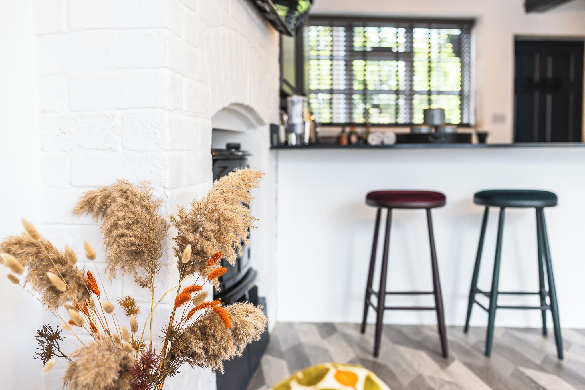 A modern kitchen with a white brick wall, a window with blinds, and two bar stools.
