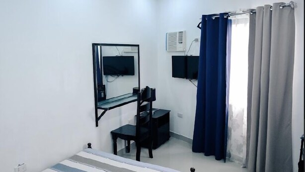 A small, sparsely furnished room with a desk, chair, and a mirror. There is a window with curtains, and an air conditioning unit mounted on the wall.