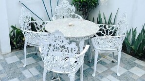 A white outdoor patio with a round table and white chairs, a barred window, and a wall-mounted clock.