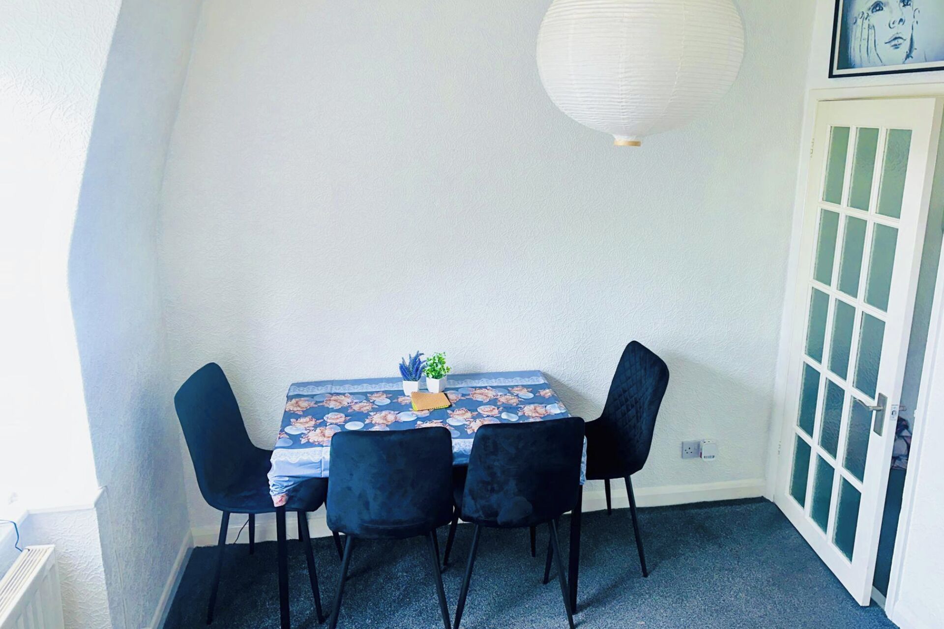 En liten matplats med ett bord dukat för fyra personer, med ett blommigt bordsduksöverdrag, två svarta stolar och en vit papperslykta som hänger ovanför.