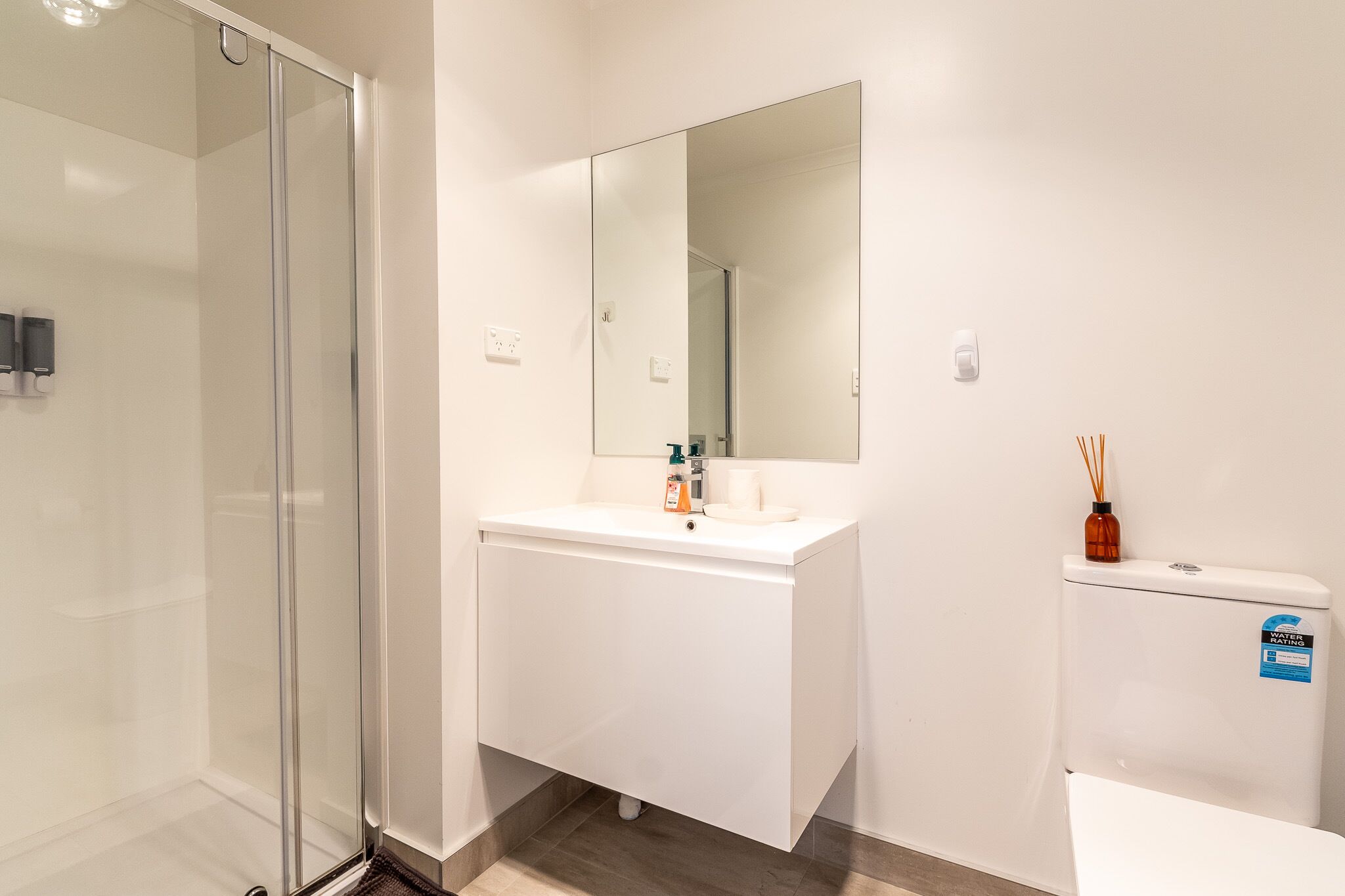 A modern bathroom with a glass shower enclosure, a white vanity with a mirror, and a toilet with a water conservation sticker.