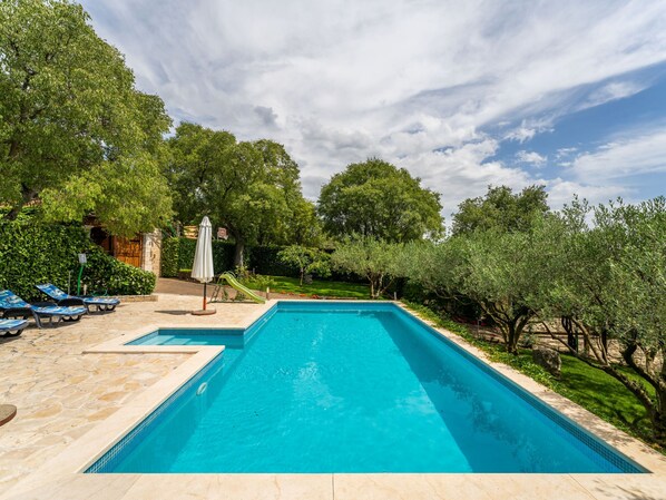 Una piscina rectangular con borde de azulejos, rodeada por una terraza de piedra. Hay sillas de descanso y una sombrilla en la terraza.