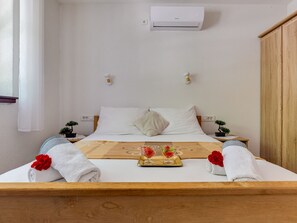 Una cama bien tendida con sábanas blancas, cabecera de madera y una bandeja con flores rojas. Hay una mesita con una planta y un aire acondicionado de pared sobre la cama.