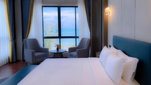 Ett hotellrum med en stor säng, två fåtöljer, ett litet bord och utsikt över havet genom stora fönster.