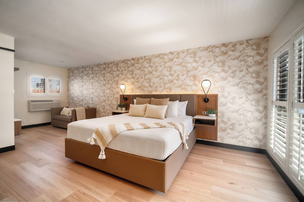 Un dormitorio moderno con una cama grande, mesitas de noche, un mueble auxiliar y un sofá, además de una ventana con persianas.