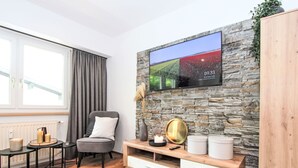 A modern living room with a stone wall, a flat-screen TV, a grey armchair, a wooden coffee table, and a white cabinet. - Am Birkenhain by Interhome (Seefeld in Tirol)