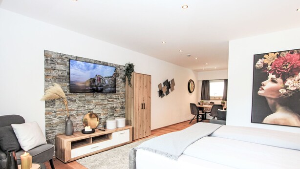A modern living room with a stone wall, a flat-screen TV, a dining area, and a large bed. - Am Birkenhain by Interhome (Seefeld in Tirol)