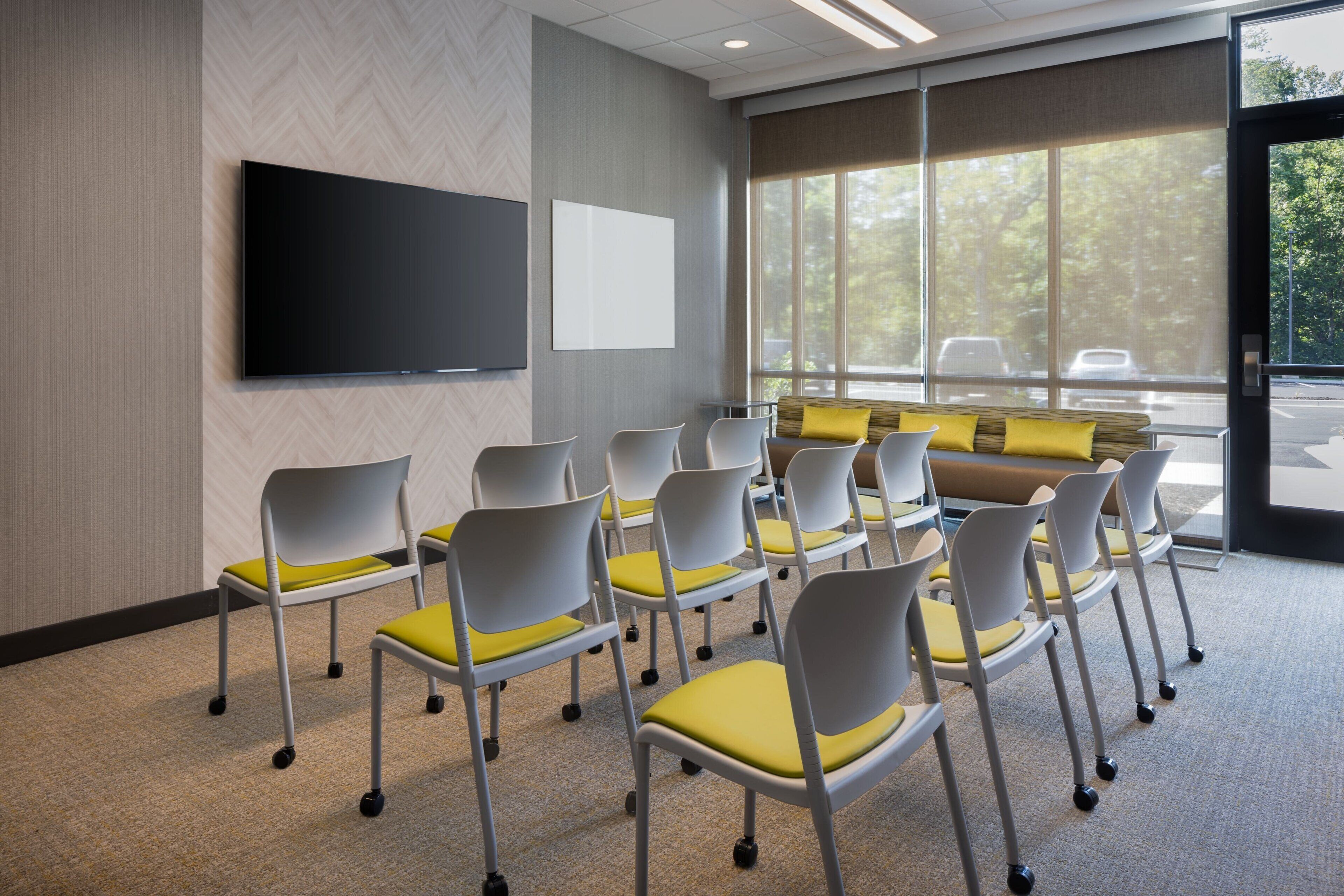 A conference room with a large flat-screen TV, white chairs with yellow cushions, a sofa with yellow cushions, and a large window with a view of trees and parked cars.