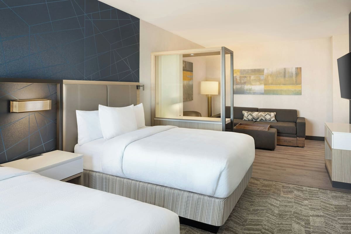 A modern hotel room with a large bed, a sofa, a desk, and a glass-enclosed shower area.