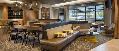 A modern hotel lobby with a seating area, a bar, and large windows offering a view of the street.