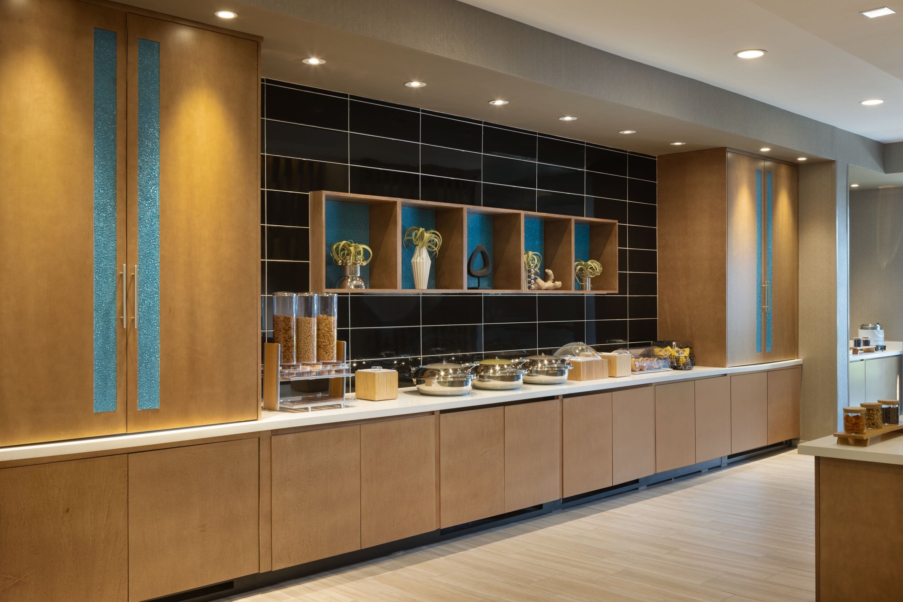 A modern kitchen with wooden cabinets, a black backsplash, and a long counter with various food items and containers.