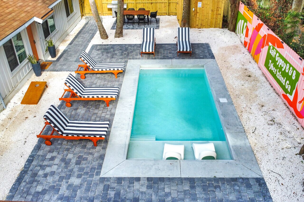A rectangular pool with lounge chairs and a table, surrounded by a paved area and a wooden fence.