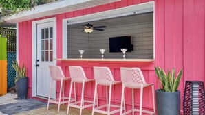 A pink outdoor bar area with four white bar stools, a ceiling fan, and a TV screen. - Salt Gypsy Unit A Expansive Outdoor Space w Pool (Tybee Island)