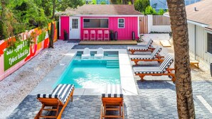 A backyard with a swimming pool, lounge chairs, and a pink building with a TV. - Salt Gypsy Unit A Expansive Outdoor Space w Pool (Tybee Island)