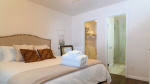 Un lit bien fait, avec une literie blanche et marron, une table de chevet avec une lampe, et une porte donnant sur une salle de bain.