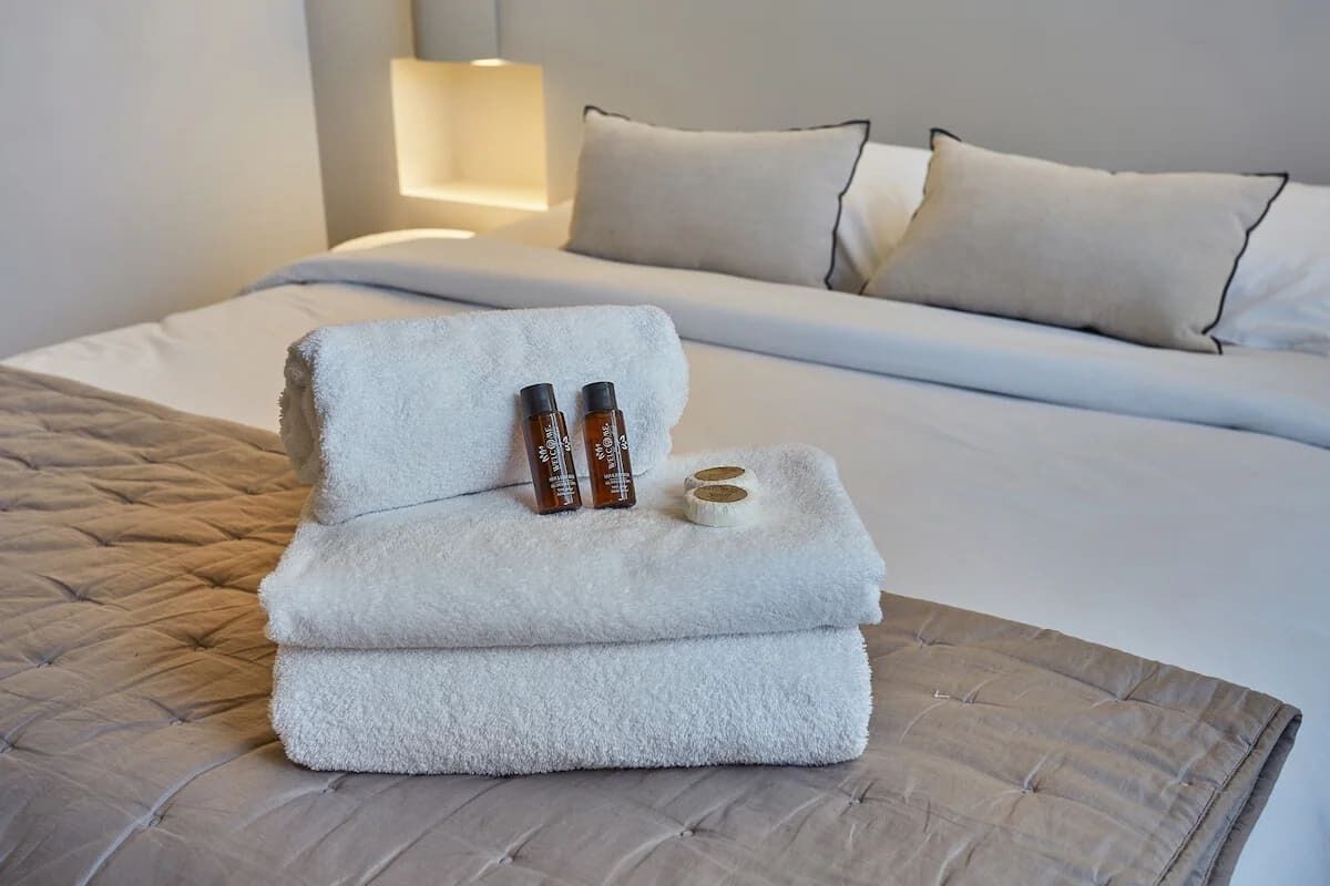 A neatly made bed with a white duvet, two bottles of lotion, and a folded towel on a tufted ottoman.