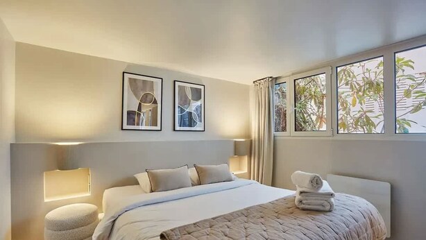 A modern hotel room with a large bed, a bedside lamp, a round ottoman, and two framed abstract artworks on the wall. - Place de la Bastille Cozy and Modern 2BD (Paris)