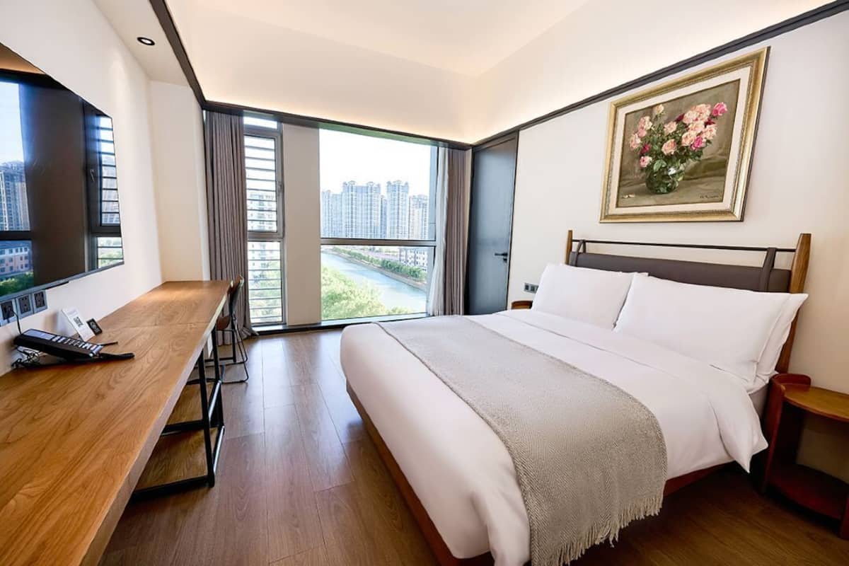 Quarto de hotel moderno com uma cama grande, uma escrivaninha com teclado, televisão e vista para a cidade pela janela.