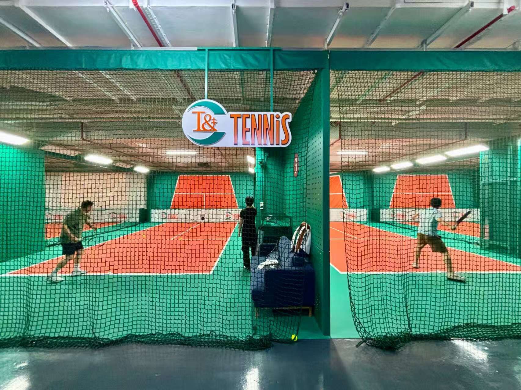 A tennis court with netting, a sign for 'J&T TENNIS', and a player in action.