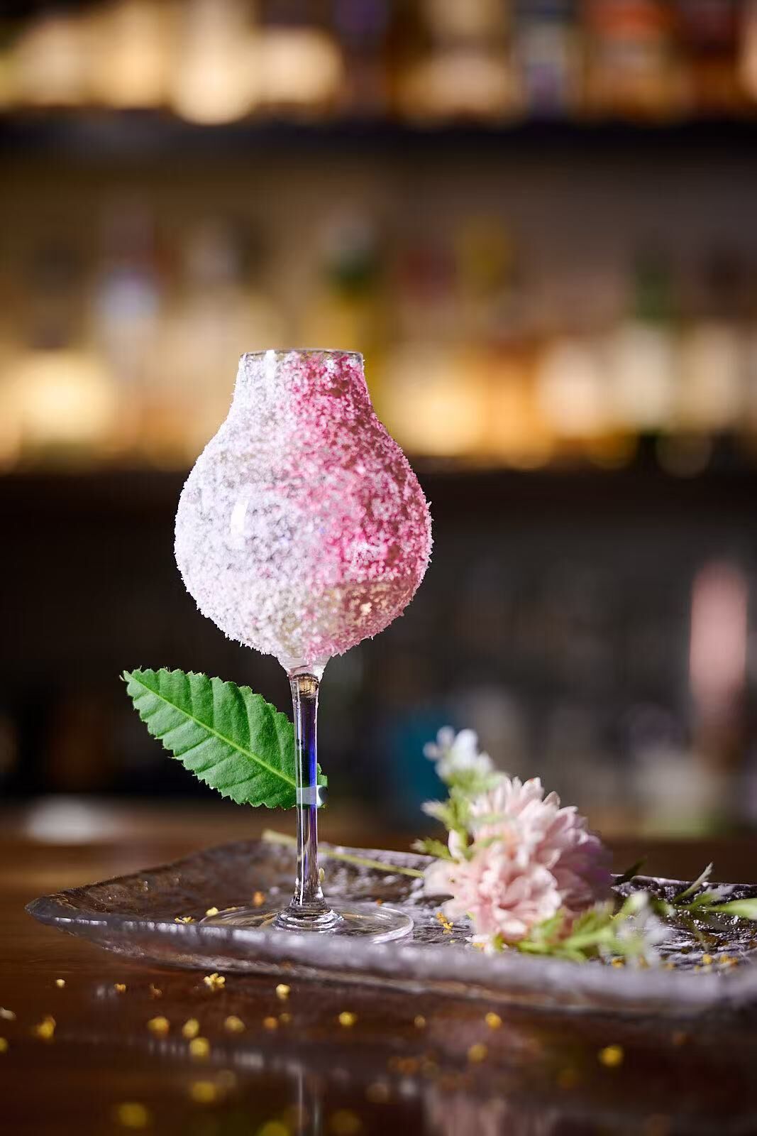 Un vaso de cóctel con aspecto helado, adornado con una hoja verde, y un fondo borroso que sugiere un bar.