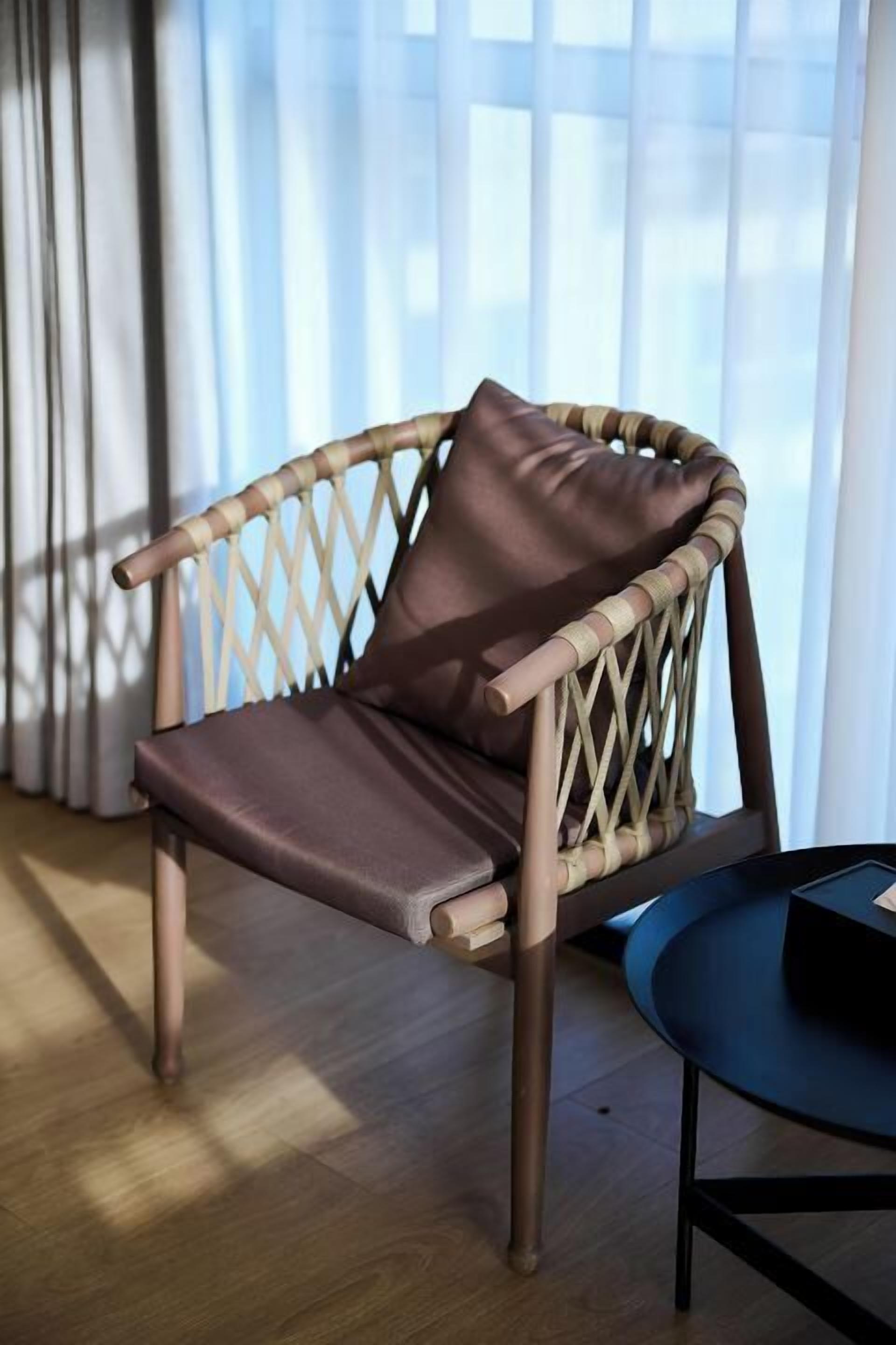 A wicker chair with a brown cushion in a room with a wooden floor and a window with sheer curtains.