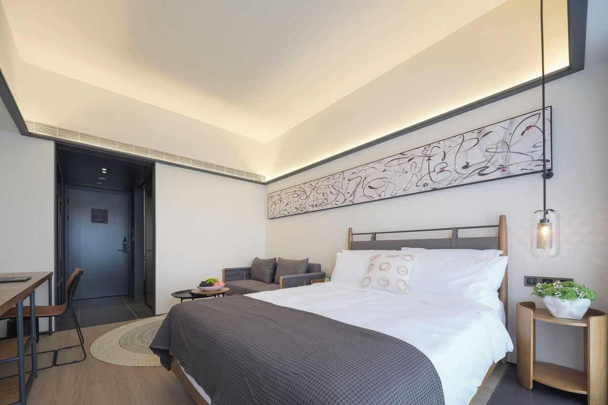 A modern hotel room with a large bed, a desk, a chair, a small table with a plant, and a wall art piece.