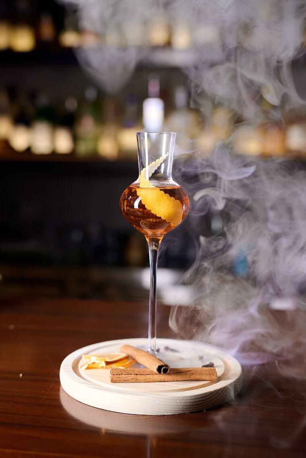 A cocktail with a lemon peel garnish on a round coaster, a cinnamon stick, and a glass of smoke on a wooden bar counter.