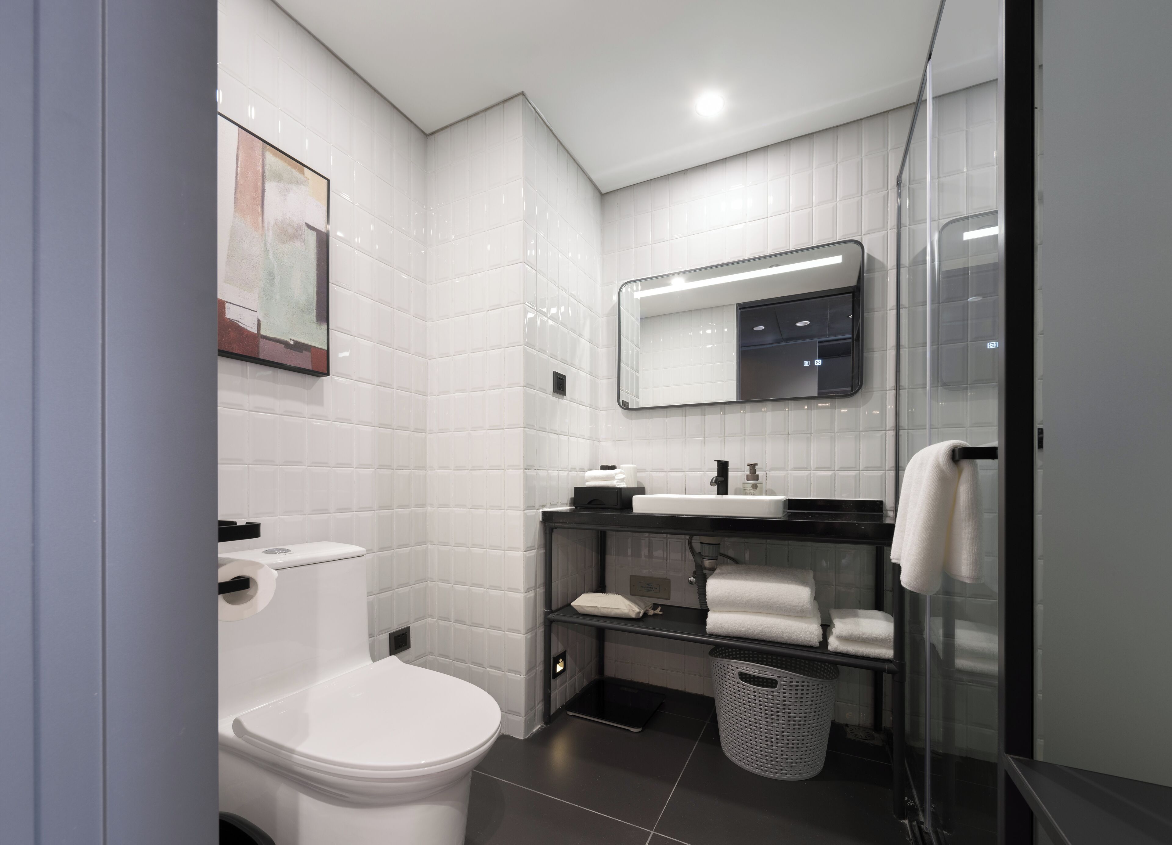 A modern bathroom with white tiles, a white toilet, a black sink, a mirror, and a glass shower enclosure.