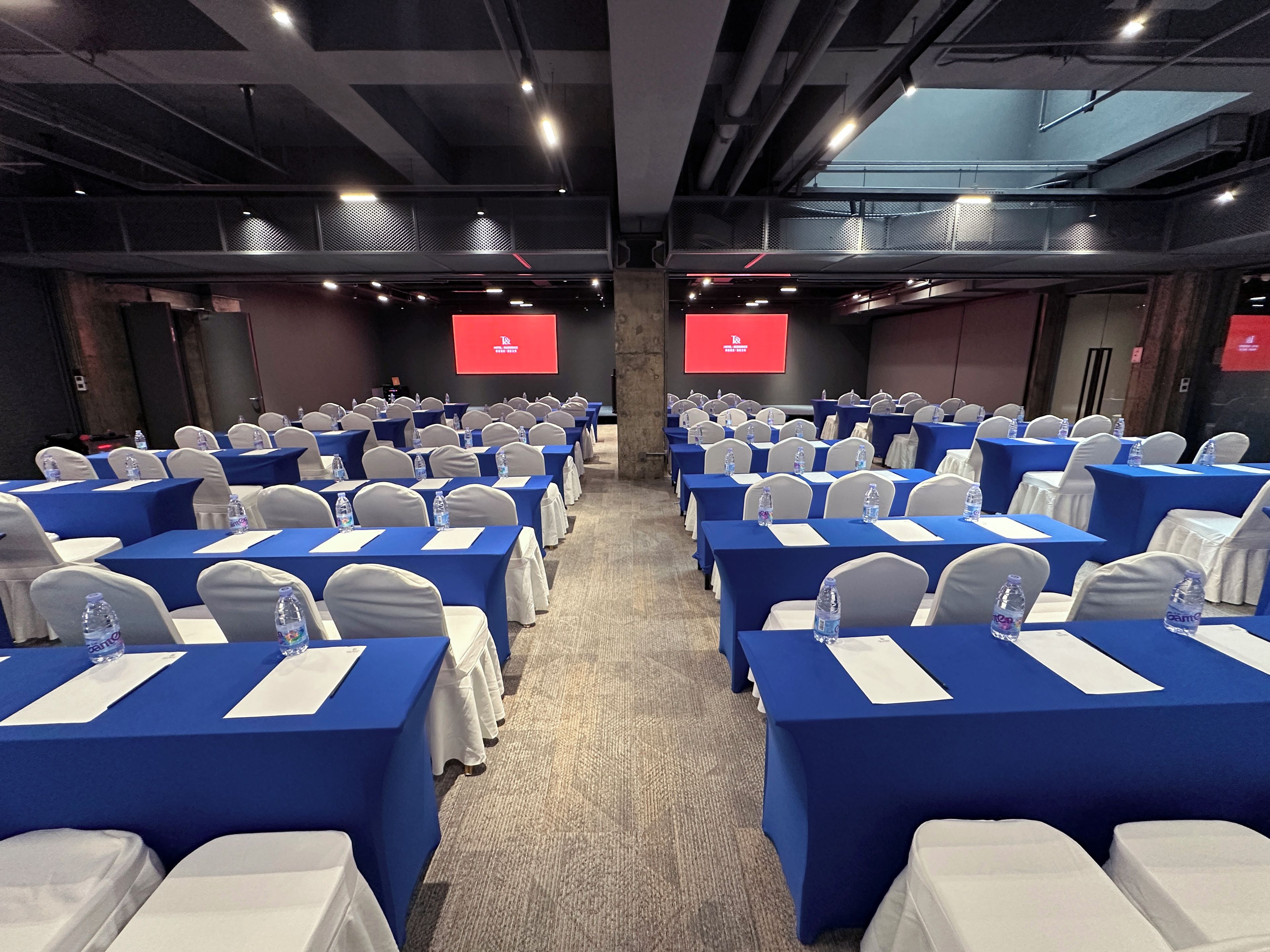 A conference room with rows of tables, chairs, and a large screen at the front.