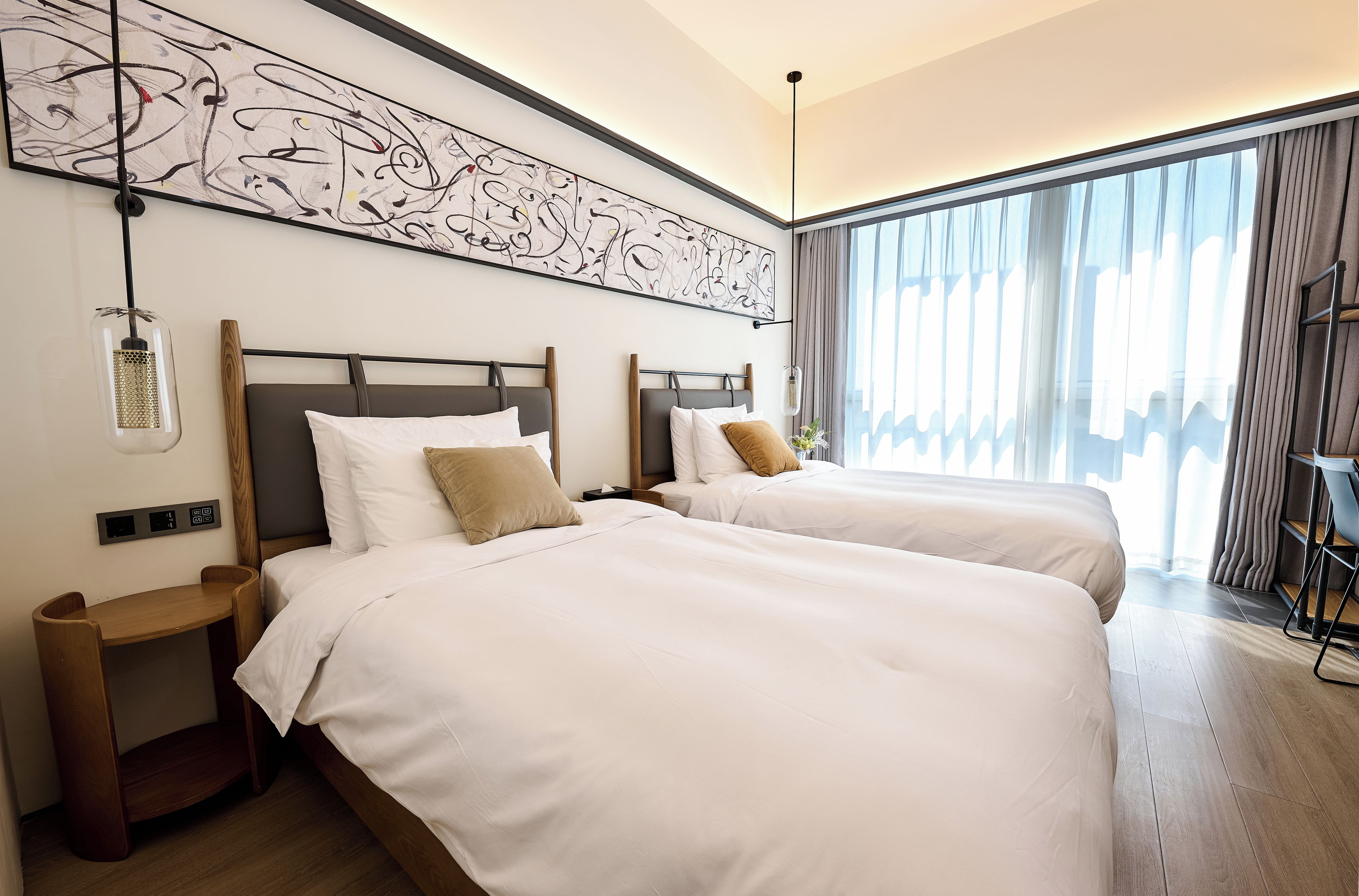 Een hotelkamer met twee bedden, een groot abstract schilderij, een moderne hanglamp en uitzicht op de stad door dunne gordijnen.