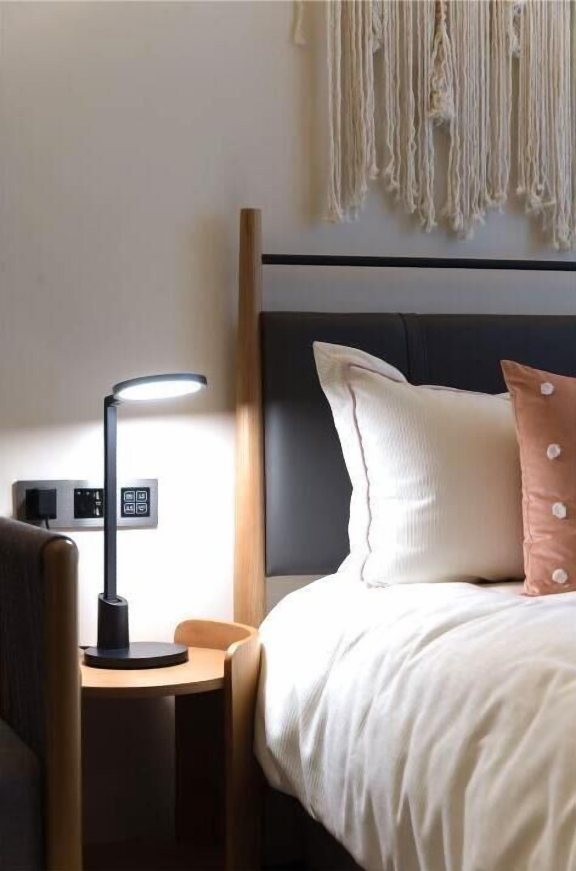 A hotel room with a bed, a bedside table with a lamp, a chair, and a wall-mounted macrame decoration.
