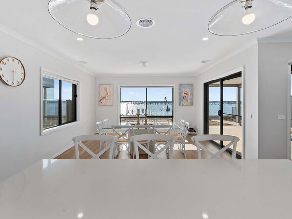 A modern dining room with a large table and chairs, large windows with a view of the sea, and a clock on the wall.