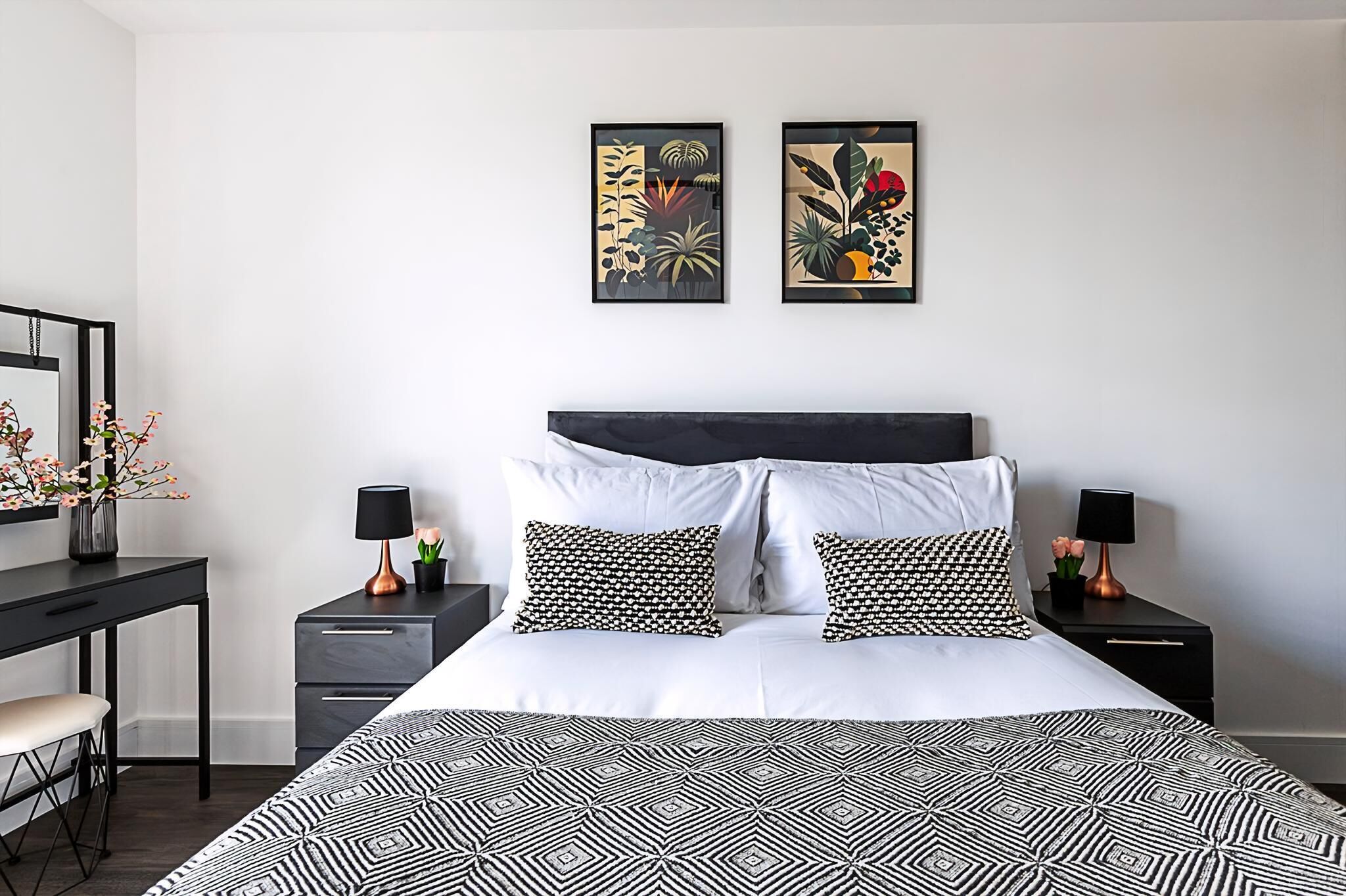 A modern bedroom with a black and white patterned bedspread, a dark headboard, two matching nightstands with lamps, and two framed botanical artworks on the wall.