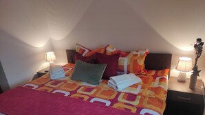 Una cama bien hecha con un edredón colorido, almohadas y toallas dobladas. Hay dos mesitas de noche con lámparas y una planta en maceta.