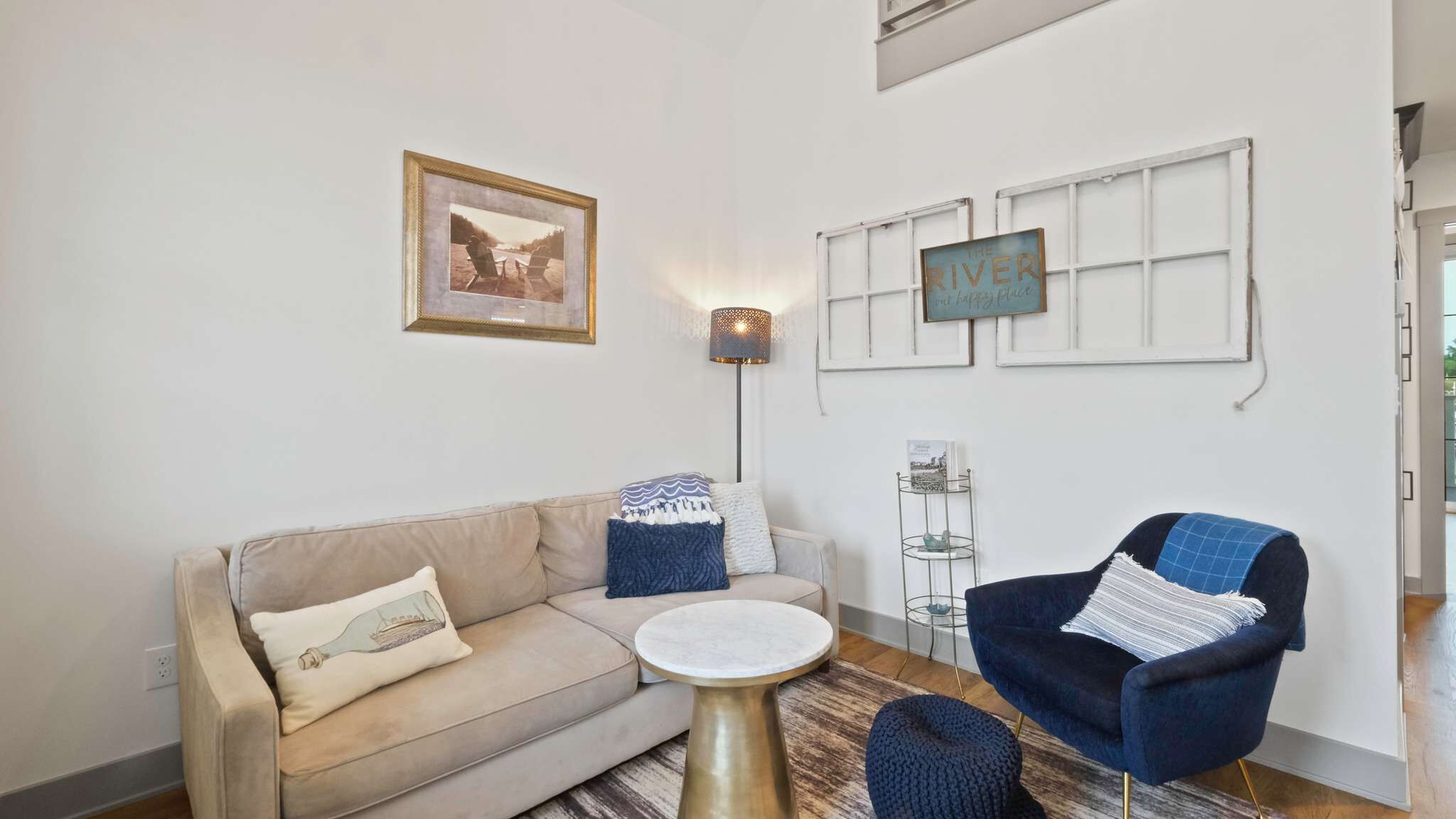 Een woonkamer met een bank, een rond bijzettafeltje, een blauwe fauteuil, een lamp en een ingelijst kunstwerk aan de muur.