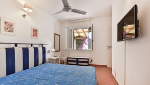 A bedroom with a bed, a ceiling fan, a window with a view of greenery, a TV mounted on the wall, and a bedside table.