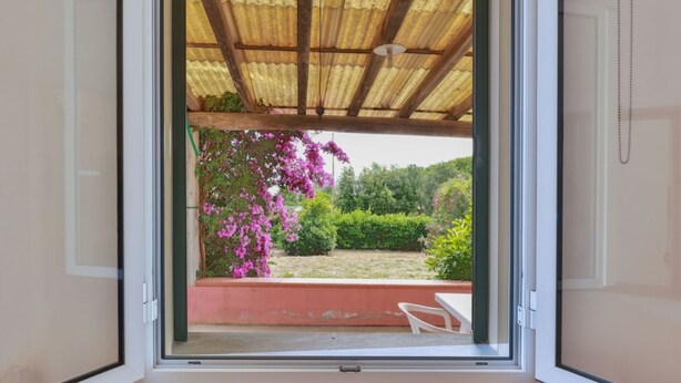 An open window with a view of a garden, a wooden pergola, and a white chair.