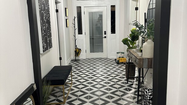 Un couloir doté d’un sol carrelé aux motifs géométriques en noir et blanc, une porte blanche au fond, et un petit banc avec une plante à côté.