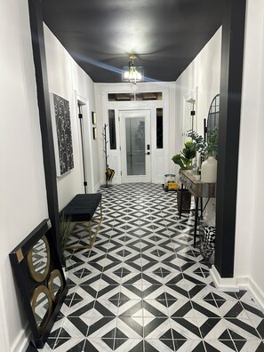 Un couloir doté d’un sol carrelé aux motifs géométriques en noir et blanc, une porte blanche au fond, et un petit banc avec une plante à côté.