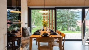 Un comedor moderno con una mesa de madera dispuesta para cuatro personas, rodeada de sillas. Un gran ventanal ofrece vistas a un paisaje boscoso.