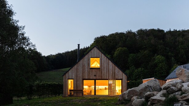 Eine Holzhütte mit großem Fenster und Schornstein, umgeben von einer grünen Wiese und Felsen.