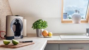 Une cuisine équipée d’une machine à café, d’une plante en pot, d’un panier de fruits et d’une fenêtre donnant sur un beau paysage.