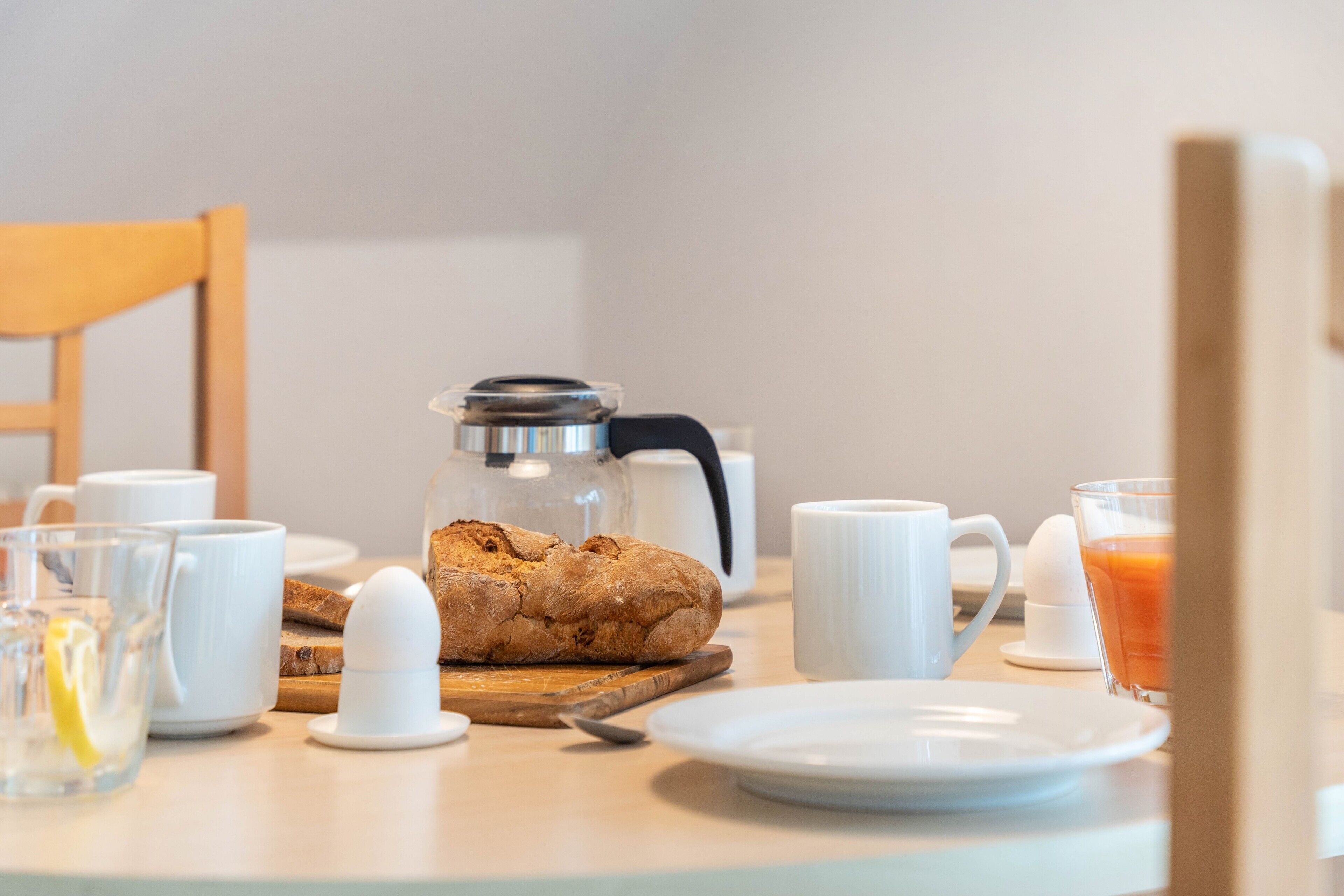 A breakfast table with a coffee pot, a loaf of bread, a glass of orange juice, and white cups.